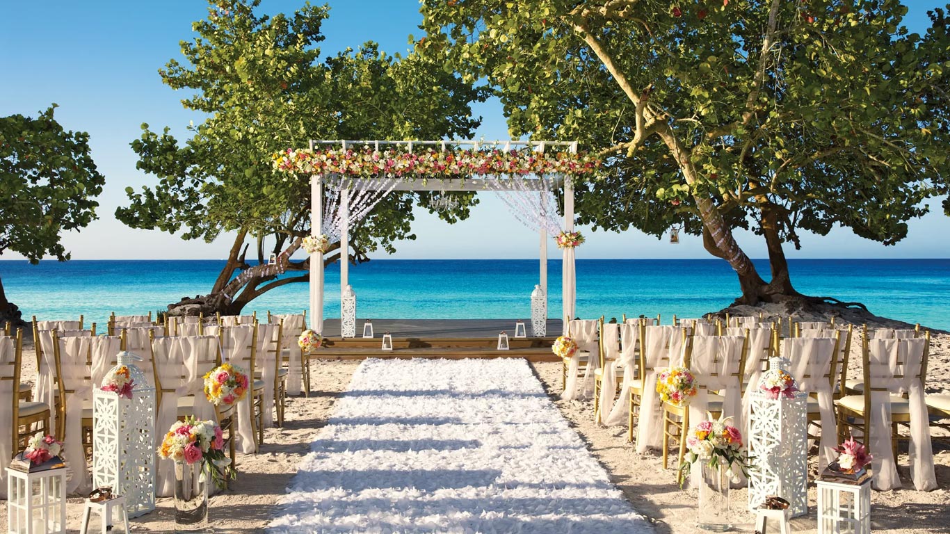 Altar de bodas frente al mar, con sillas a ambos lados de un pasillo blanco